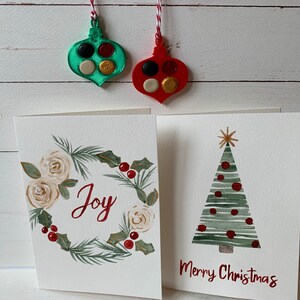 Puede incluir: Dos tarjetas de Navidad hechas a mano con ilustraciones de acuarela. Una tarjeta presenta una corona con rosas blancas y la palabra "Alegr&iacute;a" en rojo. La otra tarjeta presenta un &aacute;rbol de Navidad verde con adornos rojos y el texto "Feliz Navidad".