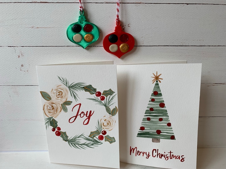 Puede incluir: Dos tarjetas de Navidad hechas a mano con ilustraciones de acuarela. Una tarjeta presenta una corona con rosas blancas y la palabra "Alegr&iacute;a" en rojo. La otra tarjeta presenta un &aacute;rbol de Navidad verde con adornos rojos y el texto "Feliz Navidad".