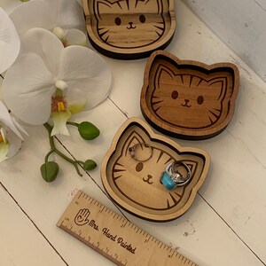 May include: Three wooden cat-shaped trinket dishes. The dishes are hand-painted with a brown wood stain and have a light brown wood color. The dishes are in the shape of a cat's face with a black outline. The dishes are on a white wooden surface.