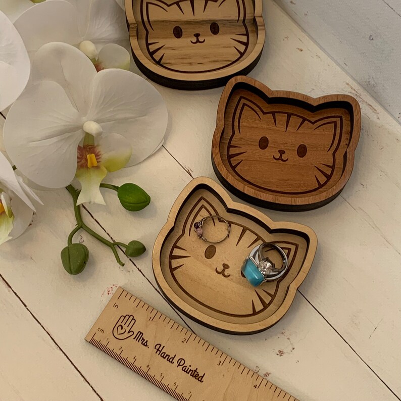 May include: Three wooden cat-shaped trinket dishes. The dishes are hand-painted with a brown wood stain and have a light brown wood color. The dishes are in the shape of a cat's face with a black outline. The dishes are on a white wooden surface.