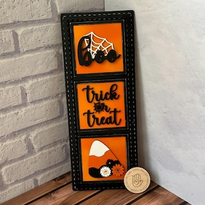 May include: Black wooden sign with three orange and black squares. The top square says "Boo" with a white spiderweb. The middle square says "trick or treat" with a small spiderweb. The bottom square has a candy corn design. The sign has a black frame with white dots.