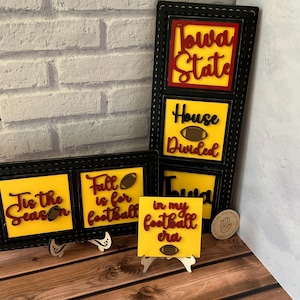 May include: Four wooden signs with yellow backgrounds and red and black lettering. The signs feature football-themed sayings: "Tis the Season", "Fall is for Football", "In my Football Era", and "Iowa State House Divided".