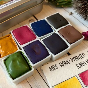 May include: A set of 8 watercolor paint pans in a metal tin. The pans are arranged in two rows of four. The colors are yellow, green, purple, blue, red, brown, black, and dark blue. The tin has a label that says "MRS. HAND PAINTED - HANDMADE IOWA PRAIRIE PA"