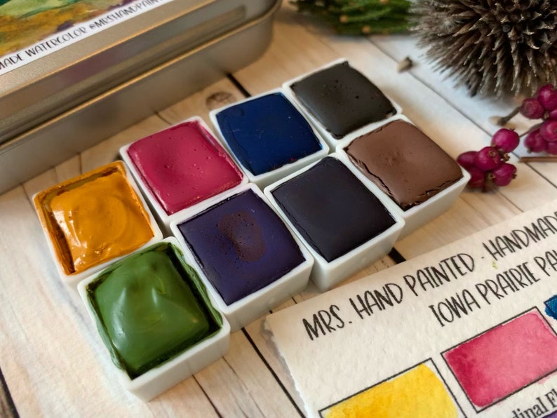 May include: A set of 8 watercolor paint pans in a metal tin. The pans are arranged in two rows of four. The colors are yellow, green, purple, blue, red, brown, black, and dark blue. The tin has a label that says "MRS. HAND PAINTED - HANDMADE IOWA PRAIRIE PA"