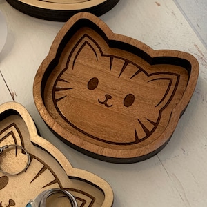 May include: A wooden trinket dish shaped like a cat's face. The dish is made of light brown wood and has a dark brown outline. The cat's face is engraved with simple features, including eyes, a nose, and whiskers.