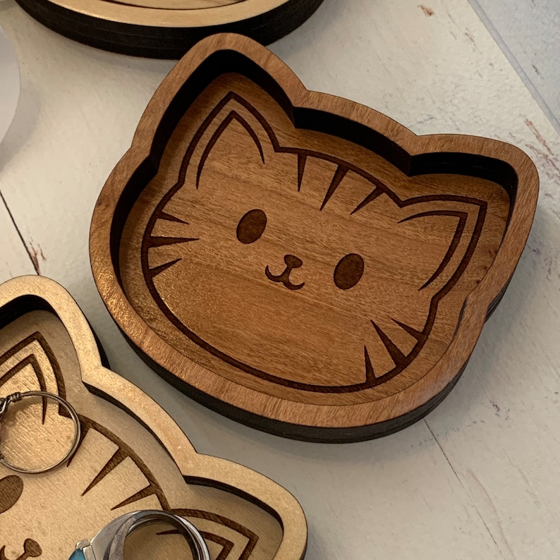 May include: A wooden trinket dish shaped like a cat's face. The dish is made of light brown wood and has a dark brown outline. The cat's face is engraved with simple features, including eyes, a nose, and whiskers.