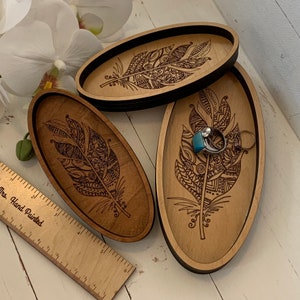 May include: Three wooden oval trays with a feather design. The trays are light brown with a dark brown border and feather engraving. One tray holds a ring with a turquoise stone and a silver ring. A wooden ruler is also visible.