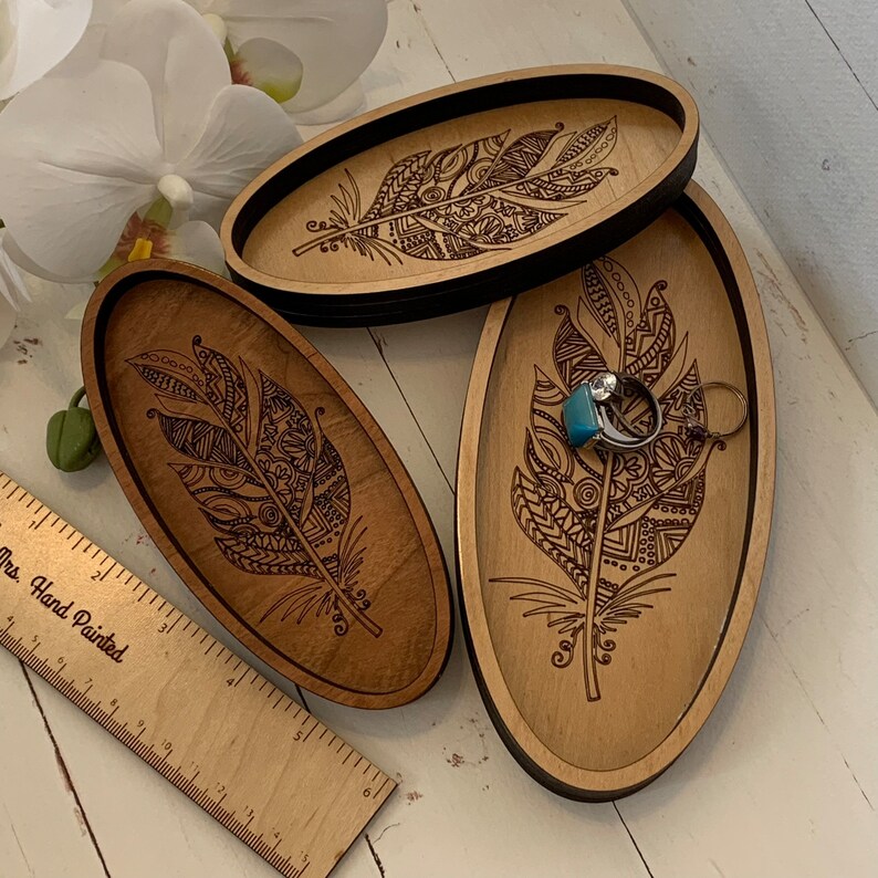 May include: Three wooden oval trays with a feather design. The trays are light brown with a dark brown border and feather engraving. One tray holds a ring with a turquoise stone and a silver ring. A wooden ruler is also visible.
