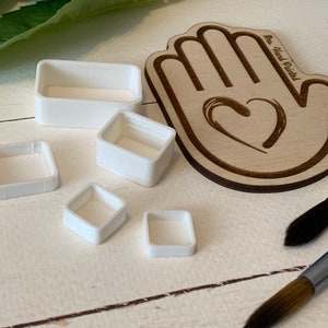 May include: Five white plastic paint palettes of different sizes. The palettes are arranged on a white wooden surface. A wooden hand-painted sign with the text "The Hand Painted" is visible in the background.