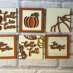 May include: Six wooden wall art pieces with a fall theme. Each square has a different design, including pumpkins, hearts, and phrases such as "Fall in Love" and "Be Thankful." The frames are orange.