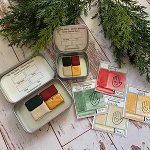 Puede incluir: Un juego de cuatro pinturas de acuarela en una lata de metal. Los colores son verde pino, crema, baya y oro. La lata tiene una etiqueta que dice "Pine, Sage, Creme - Seasonal Accents - Curtain Gourmet Palette".