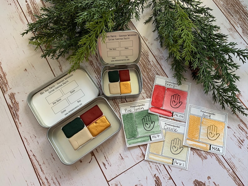 Puede incluir: Un juego de cuatro pinturas de acuarela en una lata de metal. Los colores son verde pino, crema, baya y oro. La lata tiene una etiqueta que dice "Pine, Sage, Creme - Seasonal Accents - Curtain Gourmet Palette".