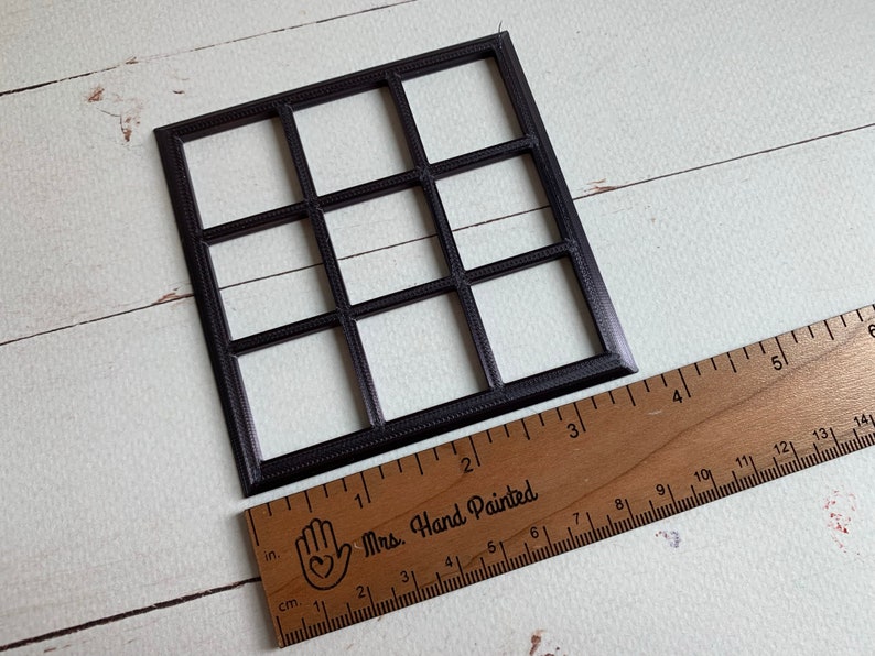 Pu&ograve; includere: Una cornice a griglia quadrata nera con nove aperture uguali, posizionata accanto a un righello di legno che mostra che la cornice misura circa 9 cm di larghezza e altezza. La cornice presenta un bordo testurizzato lungo i divisori sottili.