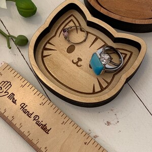 May include: A wooden cat-shaped trinket dish with a light brown finish. The dish has a carved cat face with whiskers and a small blue stone ring inside. The dish is sitting on a white surface next to a wooden ruler with the words "Mrs. Hand Painted" printed on it.