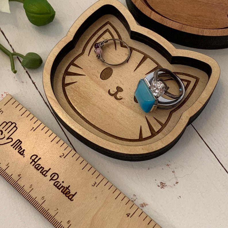 May include: A wooden cat-shaped trinket dish with a light brown finish. The dish has a carved cat face with whiskers and a small blue stone ring inside. The dish is sitting on a white surface next to a wooden ruler with the words "Mrs. Hand Painted" printed on it.