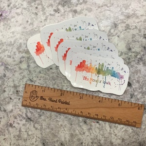 May include: A set of six rainbow watercolor stickers featuring the Des Moines, Iowa skyline. The stickers are on white paper and have a slightly irregular shape.