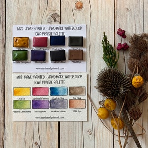 May include: A set of 10 watercolor paint pans in a cardboard box. The paint pans are labeled with the names of colors inspired by the Iowa prairie, such as "Ox Eye", "Cardinal Flower", "Prairie Sky", and "Wild Rye". The box is labeled "Mrs. Hand Painted - Handmade Watercolor Iowa Prairie Palette".