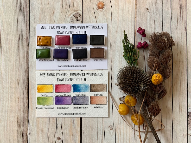 May include: A set of 10 watercolor paint pans in a cardboard box. The paint pans are labeled with the names of colors inspired by the Iowa prairie, such as "Ox Eye", "Cardinal Flower", "Prairie Sky", and "Wild Rye". The box is labeled "Mrs. Hand Painted - Handmade Watercolor Iowa Prairie Palette".