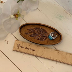 May include: A wooden, oval-shaped trinket tray with a feather design. The tray is brown with a black border and holds two rings. A wooden ruler with inch measurements is next to the tray. The tray is approximately 8 inches long.