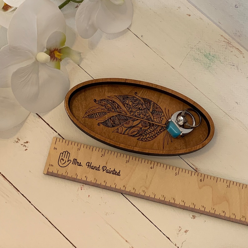 May include: A wooden, oval-shaped trinket tray with a feather design. The tray is brown with a black border and holds two rings. A wooden ruler with inch measurements is next to the tray. The tray is approximately 8 inches long.
