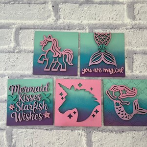 May include: Five square wooden wall art pieces with mermaid and unicorn themes. Each piece features a gradient background of teal, purple, and pink. Designs include a unicorn, mermaid tail, mermaid, and text that reads "you are magical" and "Mermaid Kisses Starfish Wishes."