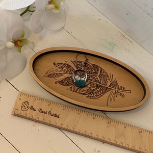May include: Oval wooden tray with a laser-etched feather design. A silver ring with a large gemstone rests on a teal fabric inside the tray. A wooden ruler with the text "Mrs. Hand Painted" is in the foreground.