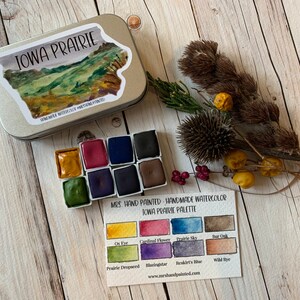 May include: A set of handmade watercolor paints in a tin with a label that says "Iowa Prairie Palette". The palette includes 10 colors: Ox Eye, Cardinal Flower, Prairie Sky, Bar Oak, Prairie Dropseed, Blazingstar, Reskirt's Blue, Wild Rye, and two other colors.