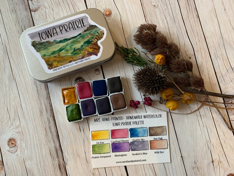 May include: A set of handmade watercolor paints in a tin with a label that says "Iowa Prairie Palette". The palette includes 10 colors: Ox Eye, Cardinal Flower, Prairie Sky, Bar Oak, Prairie Dropseed, Blazingstar, Reskirt's Blue, Wild Rye, and two other colors.