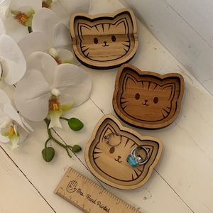 May include: Three wooden cat-shaped trinket dishes. The dishes are made of light brown wood and have a dark brown cat face engraved on them. The dishes are perfect for holding small items like rings, earrings, or other jewelry.