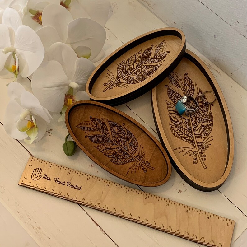 May include: Three oval wooden trays with a laser-etched feather design. The trays are in varying shades of brown and black. One tray holds rings. A wooden ruler with the text "Mrs. Hand Painted" is also present.