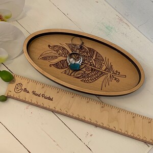 May include: Oval wooden tray with a laser-etched feather design and a black border. A silver ring with a large stone rests on a teal-colored block inside the tray. A wooden ruler with the text "Mrs. Hand Painted" is in the foreground.