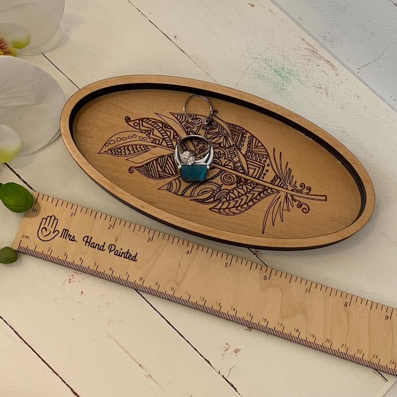 May include: Oval wooden tray with a laser-etched feather design and a black border. A silver ring with a large stone rests on a teal-colored block inside the tray. A wooden ruler with the text "Mrs. Hand Painted" is in the foreground.