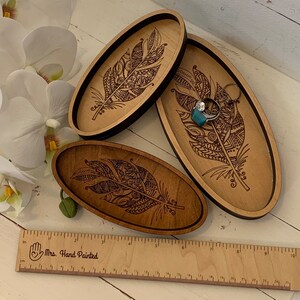 May include: Three wooden oval trays with a feather design. The trays are in varying shades of brown, with a dark border on two of them. One tray holds a ring. A wooden ruler with inch measurements is in the foreground.