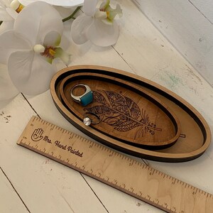 May include: Wooden oval tray set with a decorative feather design. The set includes two trays, one nested inside the other. Two rings with gemstones are inside the tray. A wooden ruler with the text "Mrs. Hand Painted" is in the foreground.