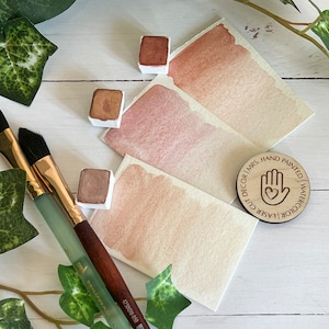 May include: Three watercolor paint pans in shades of brown, beige, and pink, with a wooden stamp that reads "Laser Cut Decor | Mrs. Hand Painted Watercolor". Two paintbrushes are also visible.