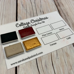 Puede incluir: Un juego de muestra de pinturas de acuarela en negro, rojo, dorado, beige y verde. Las bandejas de pintura est&aacute;n etiquetadas como "Cottage Christmas Deluxe Sample Pan Set" e incluyen los nombres de los colores: "Berry", "Pine Green", "Creme" y "Gold". La direcci&oacute;n del sitio web "www.mrs handpainted.com" est&aacute; impresa debajo de las bandejas de pintura.