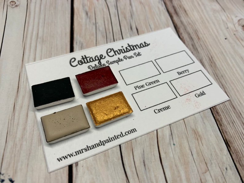 Puede incluir: Un juego de muestra de pinturas de acuarela en negro, rojo, dorado, beige y verde. Las bandejas de pintura est&aacute;n etiquetadas como "Cottage Christmas Deluxe Sample Pan Set" e incluyen los nombres de los colores: "Berry", "Pine Green", "Creme" y "Gold". La direcci&oacute;n del sitio web "www.mrs handpainted.com" est&aacute; impresa debajo de las bandejas de pintura.
