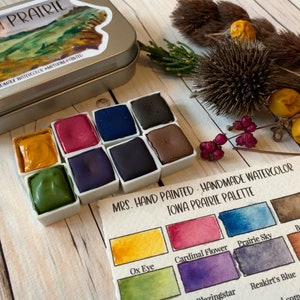 May include: A set of handmade watercolor paints in a tin with a label that reads "MRS. HAND PAINTED - HANDMADE WATERCOLOR IOWA PRAIRIE PALETTE". The palette includes colors like Ox Eye, Cardinal Flower, Prairie Sky, Bur Oak, Blazingstar, Reakirt's Blue, and Wild Rye.