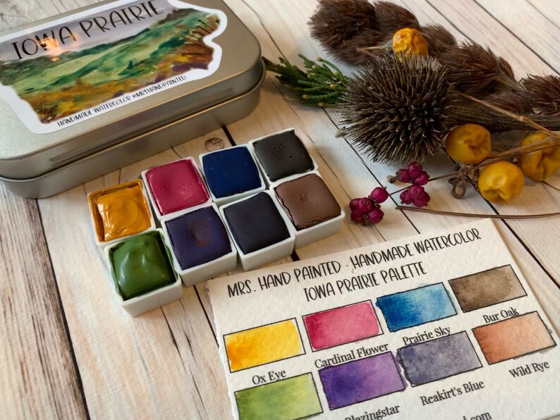 May include: A set of handmade watercolor paints in a tin with a label that reads "MRS. HAND PAINTED - HANDMADE WATERCOLOR IOWA PRAIRIE PALETTE". The palette includes colors like Ox Eye, Cardinal Flower, Prairie Sky, Bur Oak, Blazingstar, Reakirt's Blue, and Wild Rye.