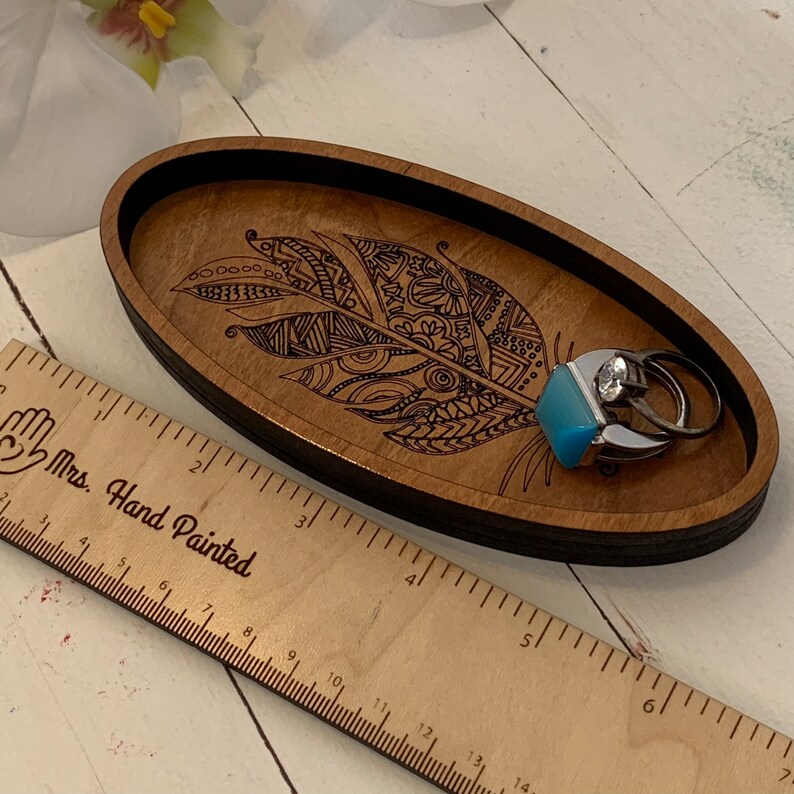 May include: Oval wooden jewelry tray with a laser-etched feather design. The tray is brown with a black rim and holds two rings, one with a blue stone and one with a clear stone. A wooden ruler is next to the tray.