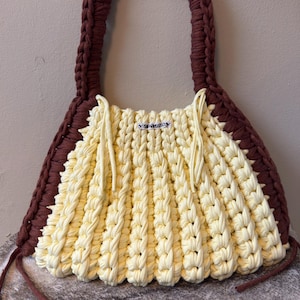May include: Handmade cream and brown crochet tote bag. The bag features a cream-colored body with a textured, vertical pattern and a brown braided handle. The bag has a rectangular shape with a label that says "Yarnessy".