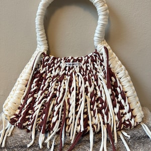May include: A handmade tote bag with a cream-colored handle and trim. The body of the bag is a woven pattern of cream and burgundy fabric with fringe. The bag has a small silver tag that reads "yorsssey".