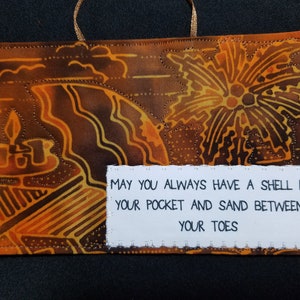 May include: A brown and orange fabric wall hanging with a white tag that reads "May you always have a shell in your pocket and sand between your toes".