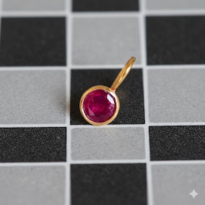 Op de afbeelding: Een gouden hanger met een ronde, helderrode robijn edelsteen. De robijn is gezet in een gouden bezel en heeft een kleine lus voor een ketting. De hanger wordt getoond tegen een zwart-wit geblokte achtergrond.