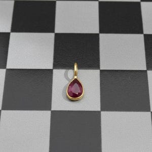 Puede incluir: Un colgante de oro con una piedra preciosa roja oscura en forma de lágrima. El colgante tiene un pequeño lazo en la parte superior para una cadena. La piedra preciosa está engastada en un bisel de oro.
