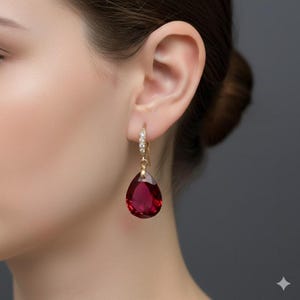 Puede incluir: Pendientes de aro dorados con un colgante de piedra preciosa roja oscura en forma de lágrima. El aro está adornado con pequeños cristales transparentes. Los pendientes se muestran en la oreja de una persona sobre un fondo gris.