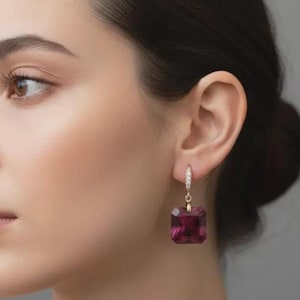 May include: Gold-tone hoop earrings with a square, deep magenta gemstone pendant. The hoops are encrusted with small, clear crystals. The earrings are shown on a person's ear, with a neutral background.