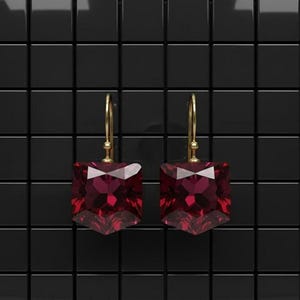 May include: A pair of gold-colored drop earrings with large, faceted, square-cut, deep red gemstones. The earrings are displayed against a black, tiled background.