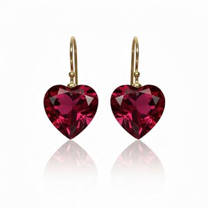 May include: A pair of heart-shaped earrings with faceted, deep red gemstones. The earrings are suspended from gold-colored hooks. The gemstones reflect light, creating a sparkling effect. The earrings are displayed on a white background.