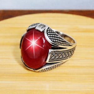 Pode incluir: Anel de prata com uma grande pedra vermelha ovalada com um efeito de estrela de seis pontas. O anel tem detalhes intrincados em prata, incluindo um padrão trançado na banda e detalhes decorativos ao redor da pedra. Sobre uma superfície de madeira.
